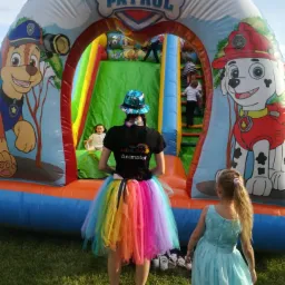 Animator w tęczowej spódnicy i błyszczącym kapeluszu stoi przed dmuchanym zamkiem z motywem Psiego Patrolu, dzieci wchodzą na zjeżdżalnię.