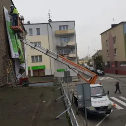 Montaż wielkoformatowej reklamy na ścianie budynku w Olesnie przy użyciu podnośnika koszowego. Pracownik w kamizelce odblaskowej mocuje baner reklamowy. Widoczna drabina oparta o ścianę.