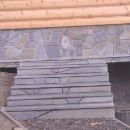 Zewnętrzne schody z szarej kamiennej okładziny prowadzące do wejścia pod budynek z drewnianą konstrukcją i kamienną elewacją, widoczne ziemia i gruz.