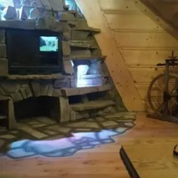 Kamienny kominek z półkami w góralskim stylu, z wbudowanym telewizorem i podświetleniem LED na podłodze, w tle drewniana ściana i kołowrotek.