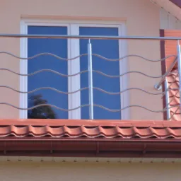 Balkon z falistą konstrukcją ze stali, odbicie nieba w szybach okiennych, widok z dołu na czerwoną dachówkę.