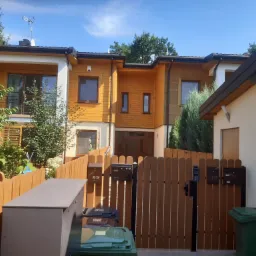 Nowoczesny dom w zabudowie szeregowej z drewnianą elewacją, widoczny zza drewnianego ogrodzenia z kilkoma skrzynkami pocztowymi i pojemnikami na śmieci.