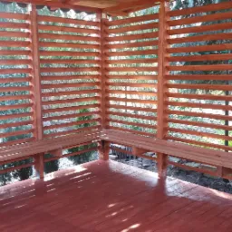 Wnętrze nowej, drewnianej altany z poziomymi listwami w ścianach i wbudowanymi ławkami, pomalowane na brązowo, z widokiem na zewnątrz.