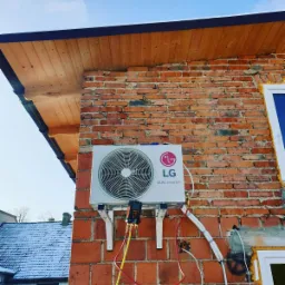 Montaż klimatyzatora LG Dual Inverter na ceglanej ścianie domu, widoczne przewody i manometry serwisowe, błękitne niebo w tle.