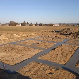 Wylane betonowe ławy fundamentowe w gruncie, przygotowane pod budowę, z widocznymi drewnianymi elementami szalunku, na tle pola i zabudowań wiejskich w oddali, przy bezchmurnym, błękitnym niebie.