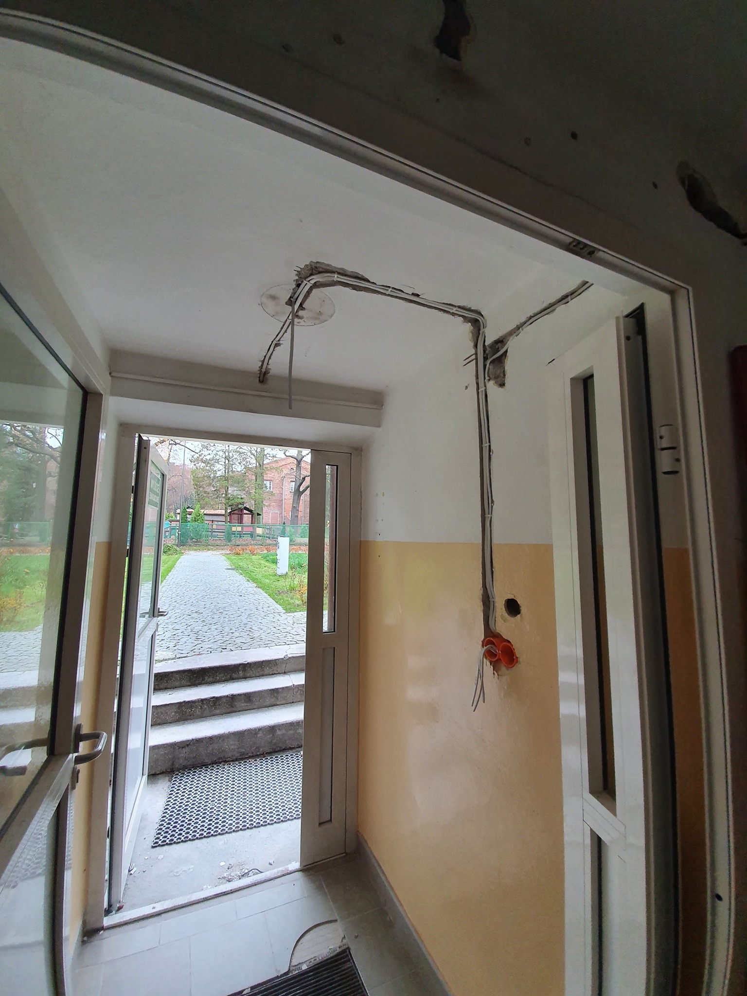 Korytarz wejściowy w budynku z widocznymi przewodami elektrycznymi prowadzonymi po ścianie i suficie, z pomarańczową puszką instalacyjną na ścianie. Przez otwarte drzwi widoczny chodnik i zieleń...