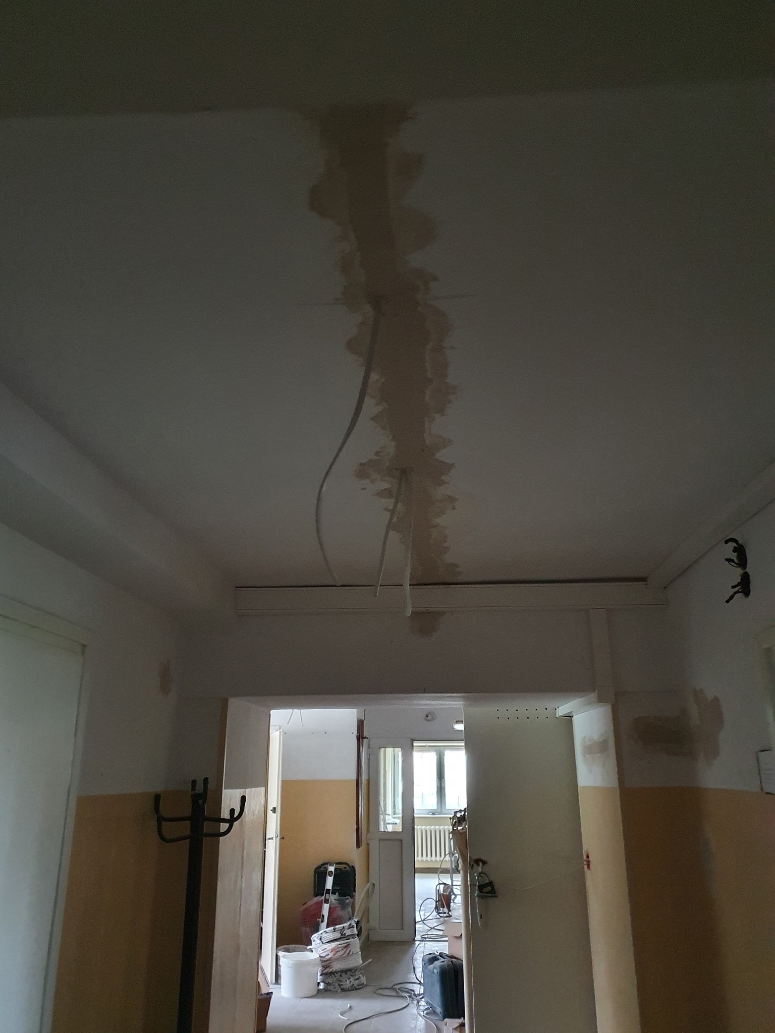 Fragment sufitu w trakcie remontu z widocznymi przewodami elektrycznymi, śladami po bruzdach i częściowo położoną gładzią szpachlową. W tle widoczne pomieszczenie z narzędziami i workami z zaprawą.