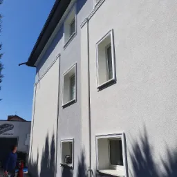 Jasnoszara elewacja budynku z białymi obramowaniami okien, widok z dołu, z pracownikiem z farbą w tle i cieniem drzewa na ścianie.