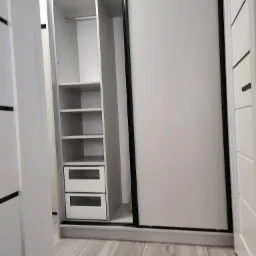 Wnętrze szafy wnękowej z przesuwnymi drzwiami, widoczna część z półkami i dwoma szufladami w kolorze białym, na podłodze panele imitujące drewno.