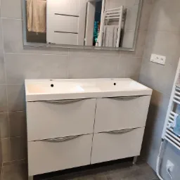 Biała szafka pod umywalkę z dwoma umywalkami i czterema szufladami, chromowane uchwyty, lustro z podświetleniem LED, szare płytki na ścianach i podłodze, grzejnik drabinkowy z ręcznikiem.