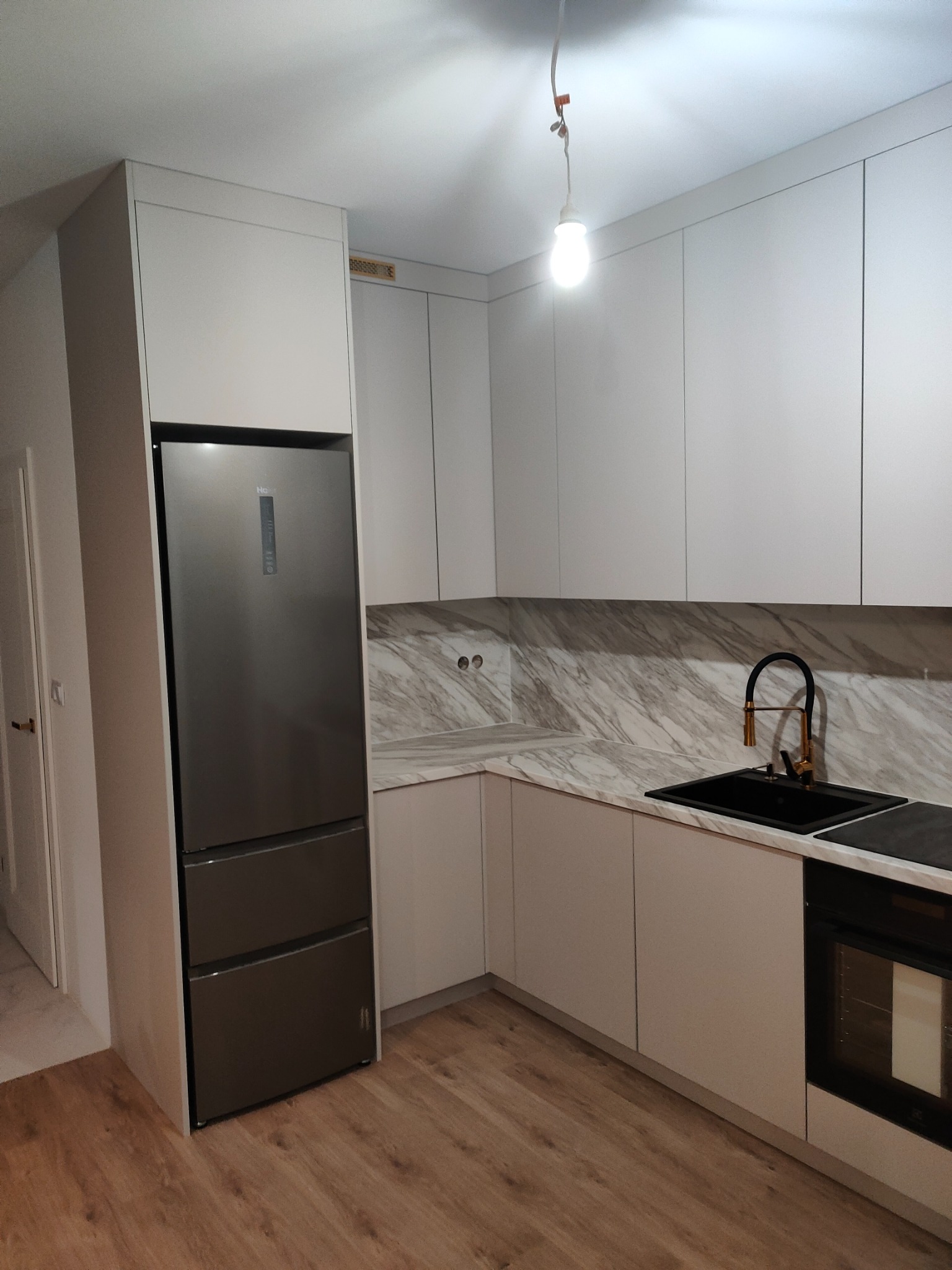 Nowoczesna kuchnia z szarymi frontami, marmurowym blatem, czarną armaturą i lodówką w zabudowie, oświetlona wiszącą żarówką