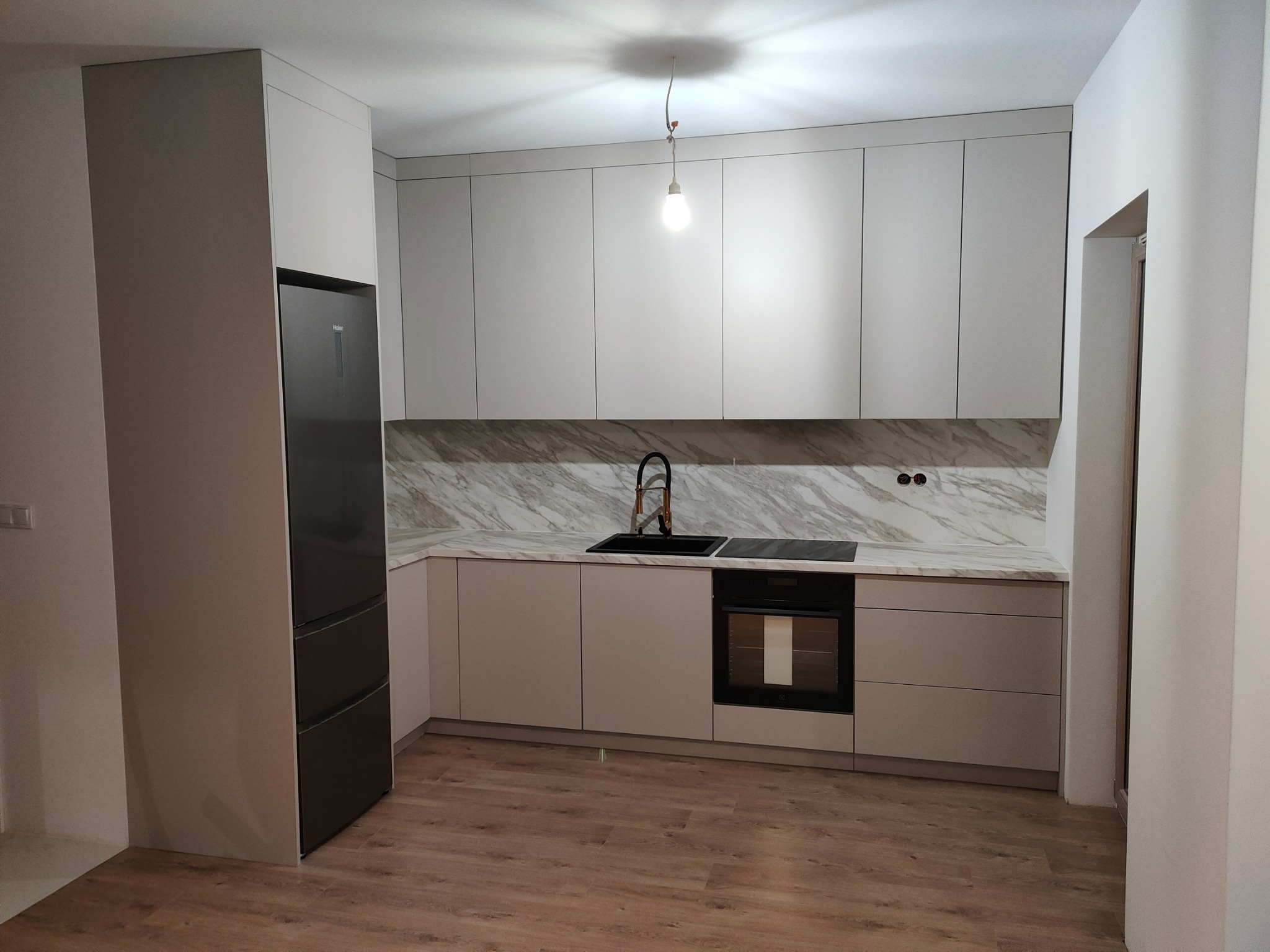 Nowoczesna kuchnia z jasnymi, gładkimi frontami szafek, marmurowym blatem i czarną armaturą. Widoczny piekarnik i lodówka w zabudowie. Uwagę zwraca surowa żarówka zwisająca z sufitu.