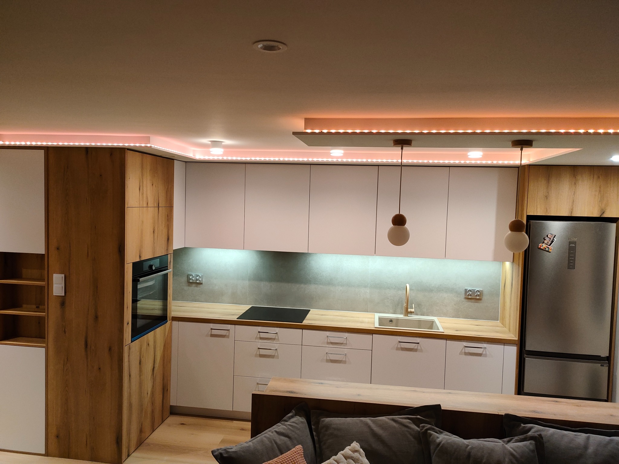Nowoczesna kuchnia z białymi szafkami, drewnianymi akcentami i oświetleniem LED podszafkowym oraz sufitowym, widziana z perspektywy części wypoczynkowej z szarą sofą.