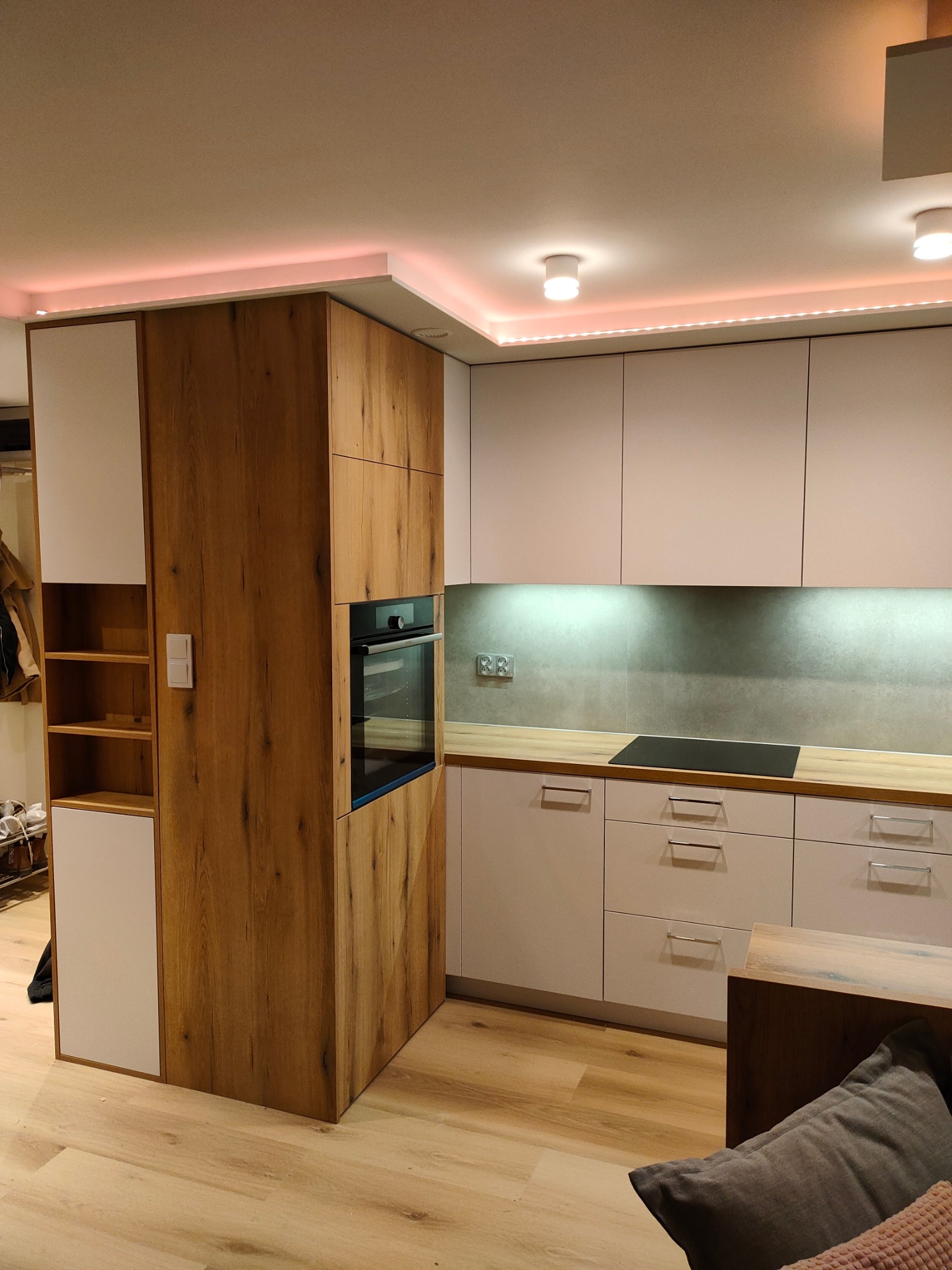 Nowoczesna kuchnia: białe szafki z drewnianym blatem, drewniana zabudowa z piekarnikiem i półkami, oświetlenie LED podszafkowe i sufitowe.