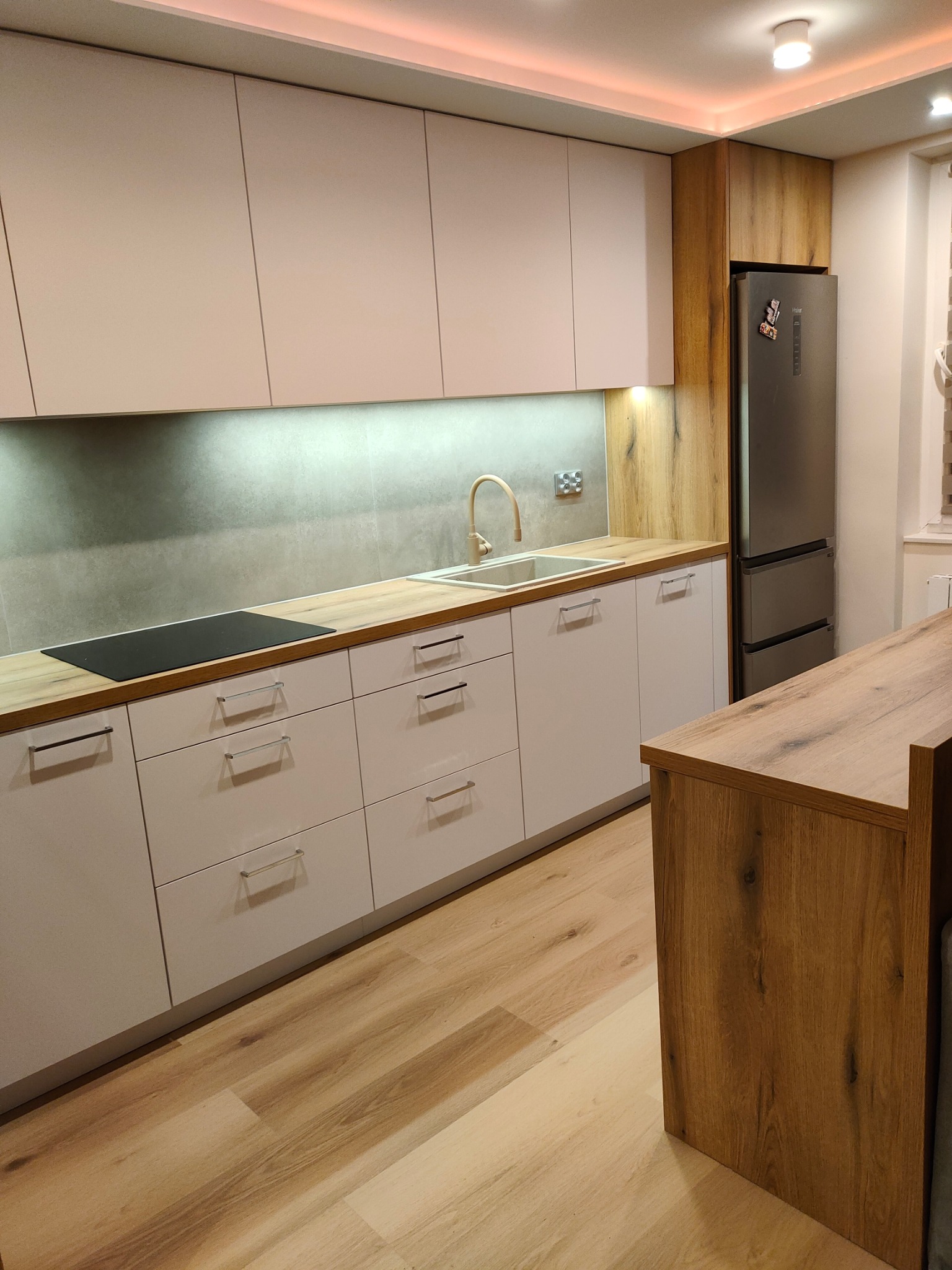 Jasna kuchnia z drewnianymi akcentami: białe szafki z podłużnymi uchwytami, drewniany blat, szary panel nad blatem, drewniana zabudowa lodówki, oświetlenie LED podszafkowe i sufitowe,...