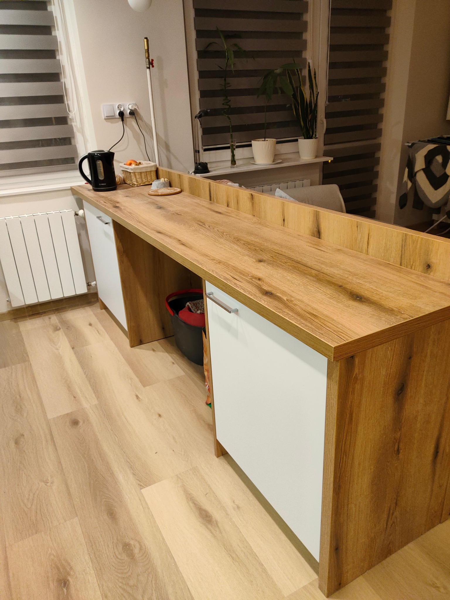 Długa, drewniana lada z białymi szafkami dolnymi w jasnym wnętrzu. Na blacie czajnik elektryczny, koszyk z owocami i podkładka. Za ladą widoczne rośliny doniczkowe i żaluzje.