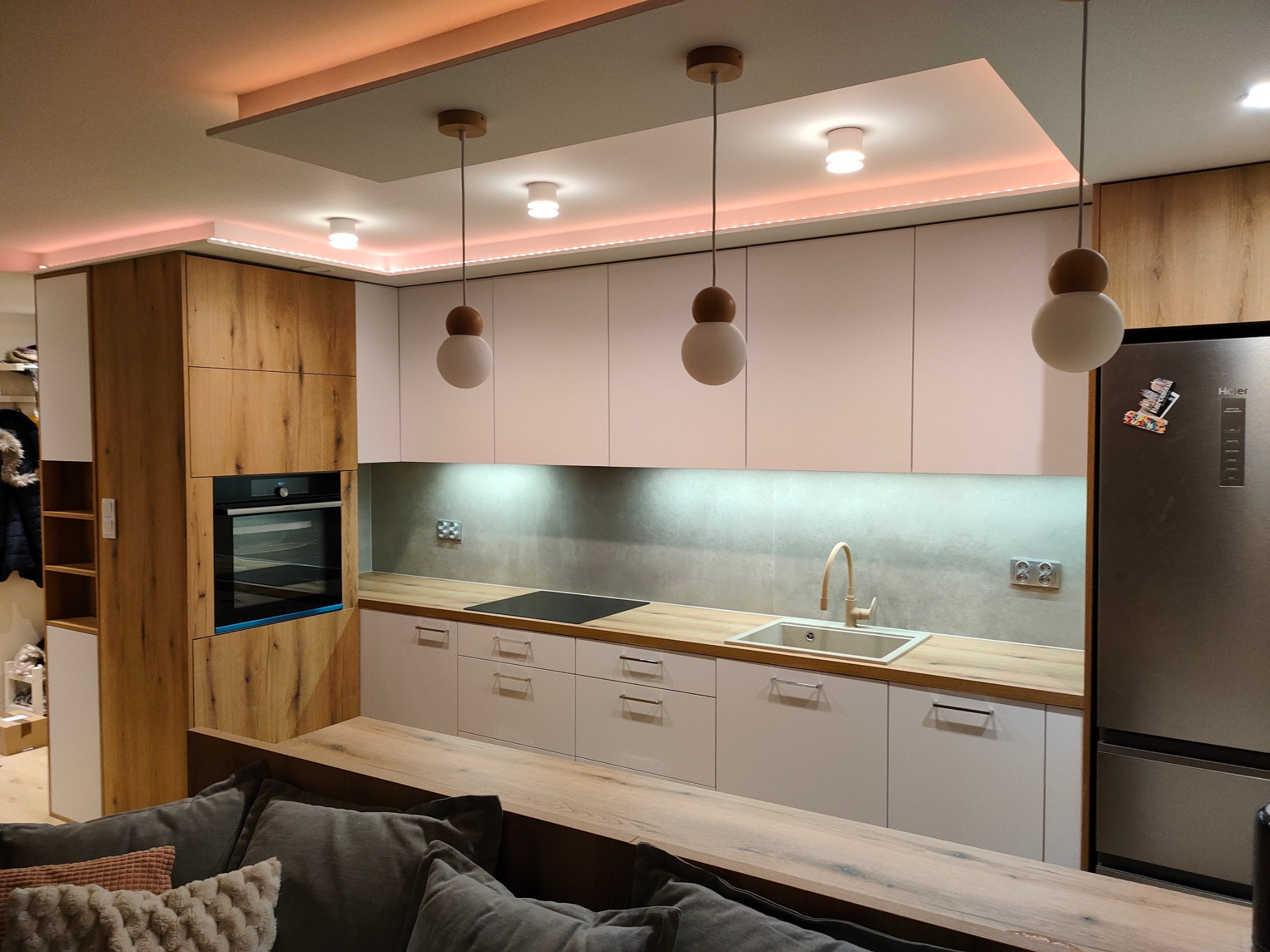 Nowoczesna kuchnia z białymi szafkami i drewnianymi akcentami, oświetlona lampami wiszącymi i listwami LED, widok z perspektywy salonu z fragmentem szarej sofy.