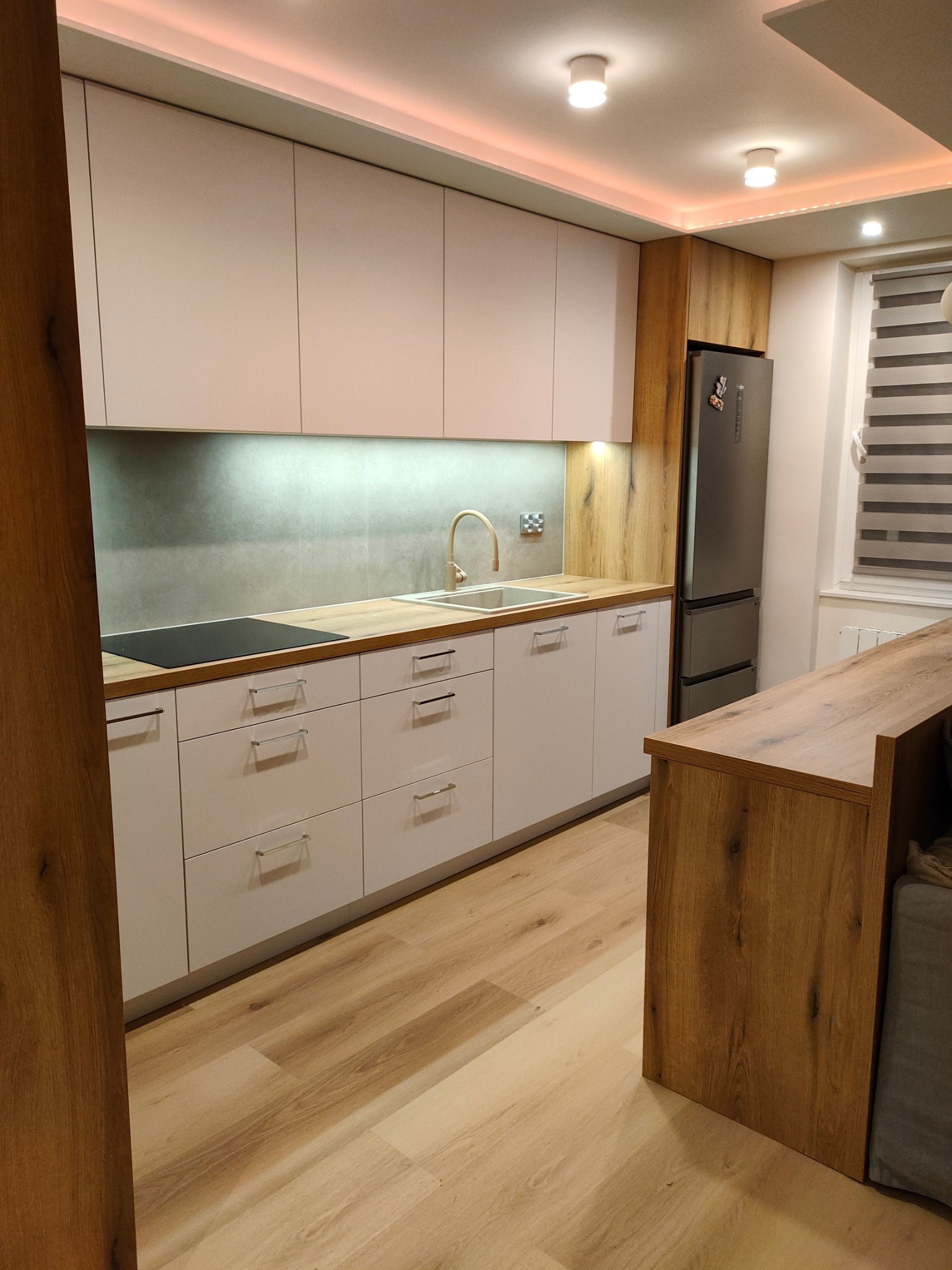 Nowoczesna kuchnia z białymi frontami, drewnianym blatem i wykończeniem w kolorze drewna, z oświetleniem LED podszafkowym i listwą LED na suficie, widoczna płyta indukcyjna i zlewozmywak z baterią...