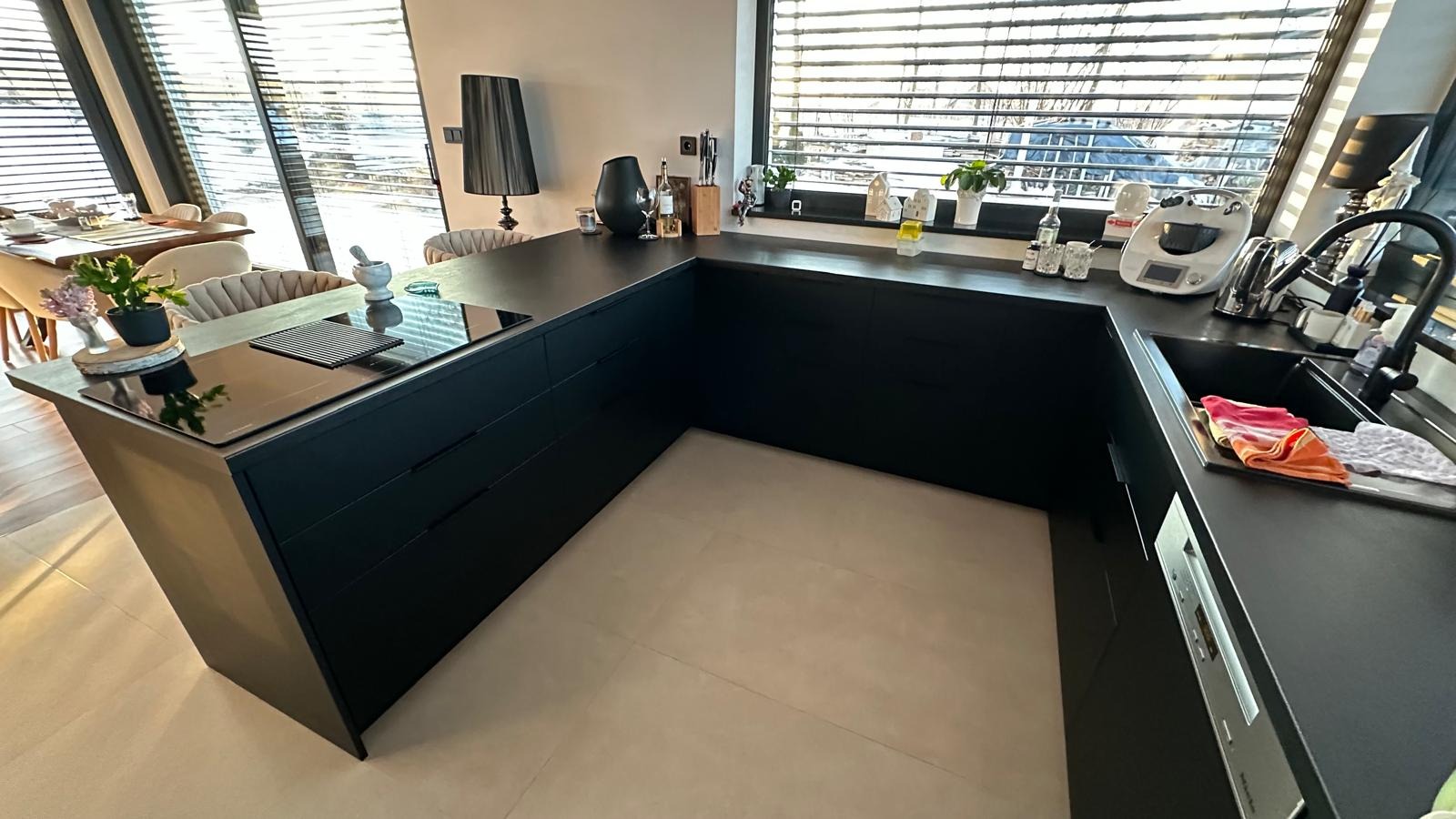 Nowoczesna, czarna kuchnia w kształcie litery L z wyspą. Minimalistyczny design, jasne płytki podłogowe, duże okno z żaluzjami. Na blacie akcesoria kuchenne i dekoracje.