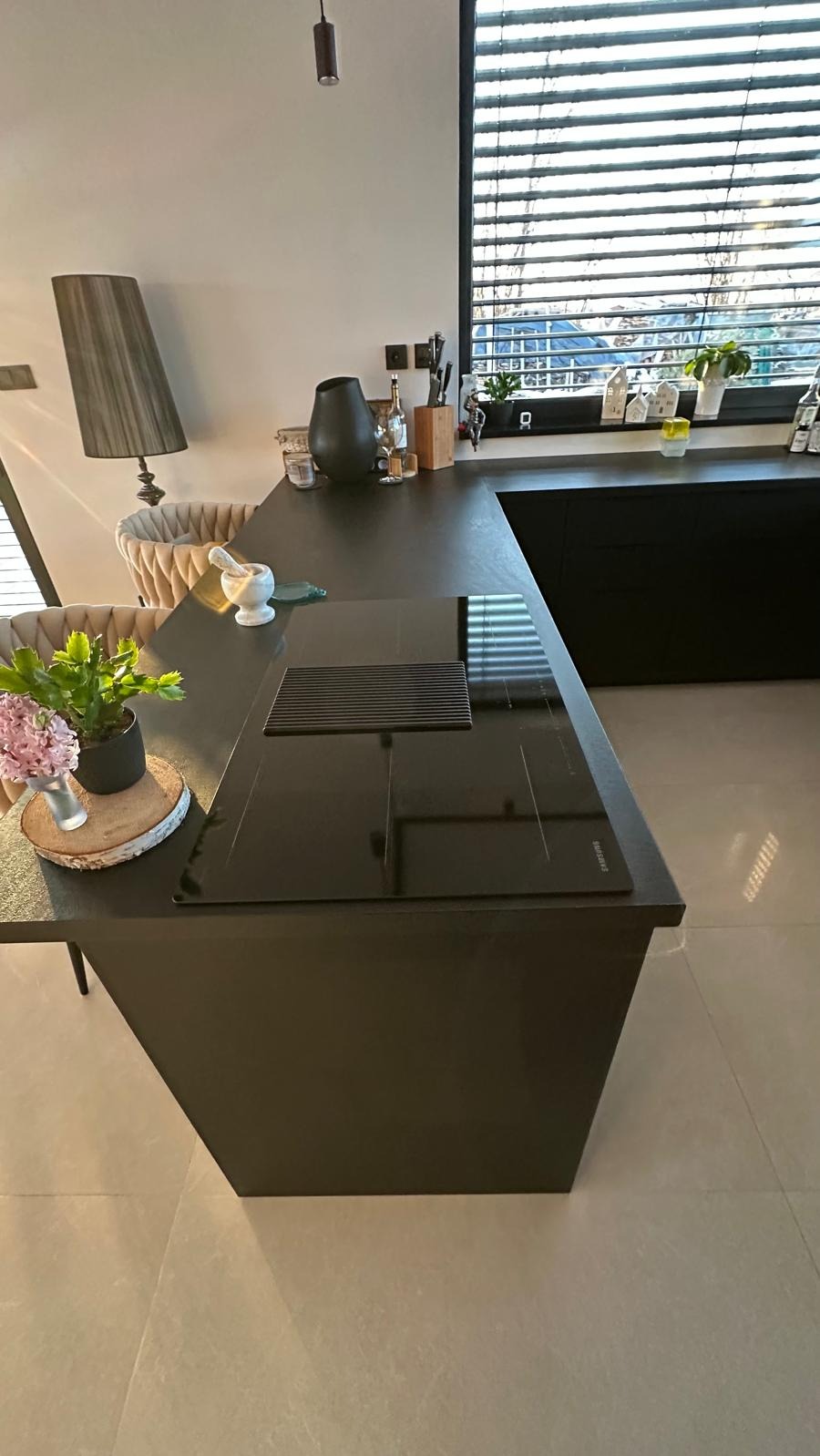 Nowoczesna, czarna wyspa kuchenna z płytą indukcyjną i pochłaniaczem w blacie. Na blacie dekoracje: wazon, moździerz, kwiaty. W tle okno z żaluzjami i lampa.