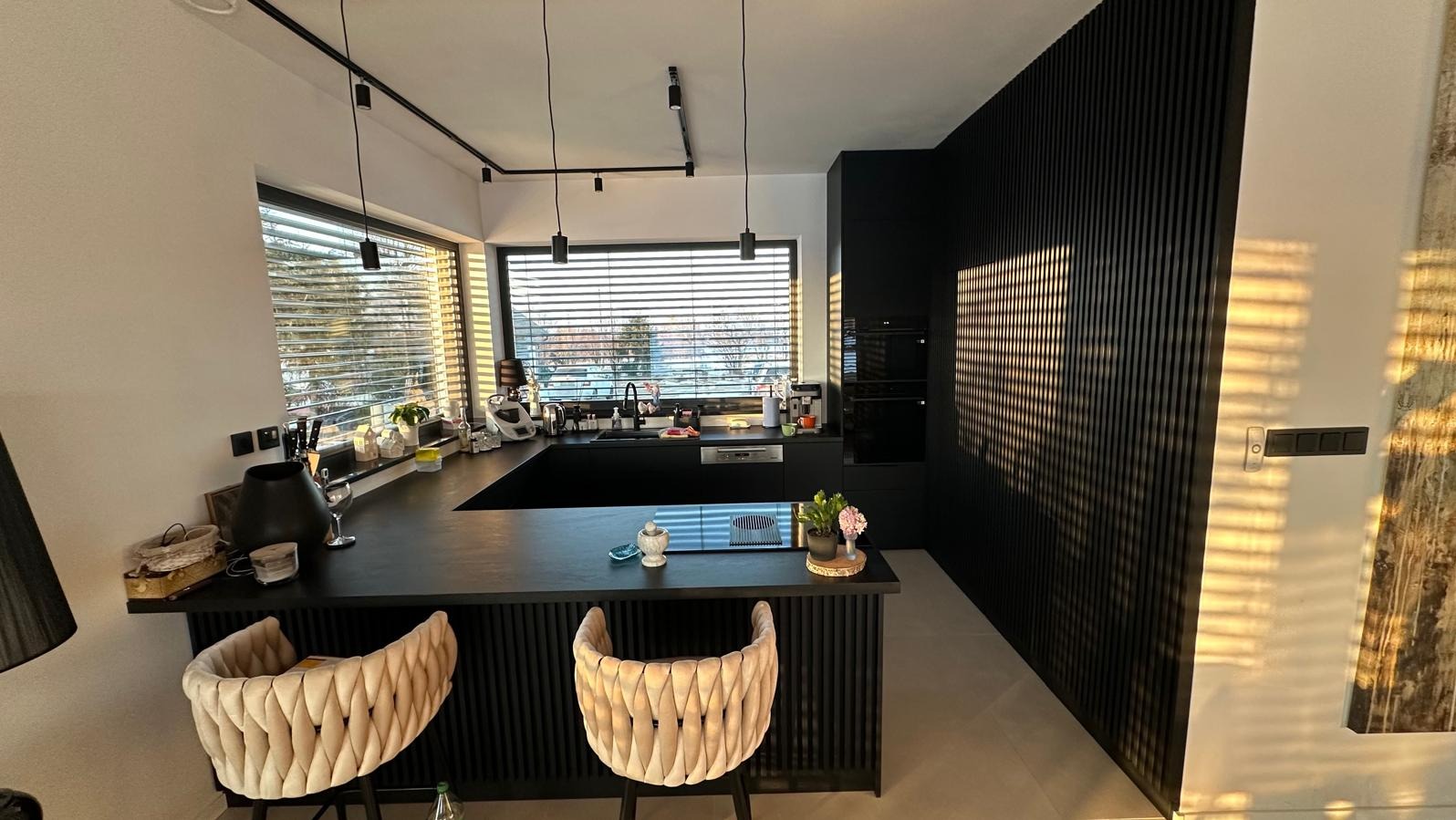 Nowoczesna, czarna kuchnia na wymiar ze ścianą z lameli w Skawinie. Widoczne krzesła barowe i okno z żaluzjami. Minimalistyczny design i oświetlenie punktowe.