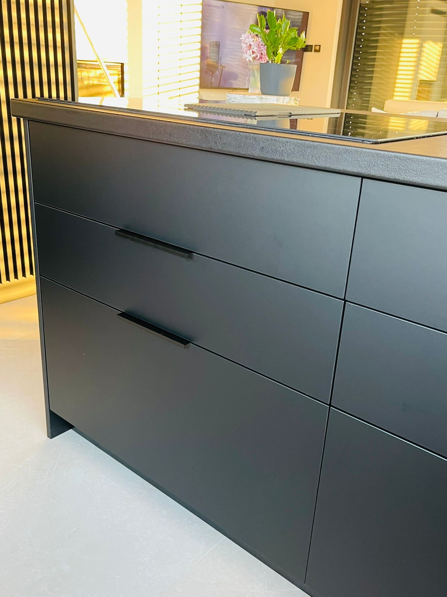 Matowa, grafitowa wyspa kuchenna z minimalistycznymi uchwytami i blatem z płyty indukcyjnej. Na blacie kwiaty w doniczce. Nowoczesny design.