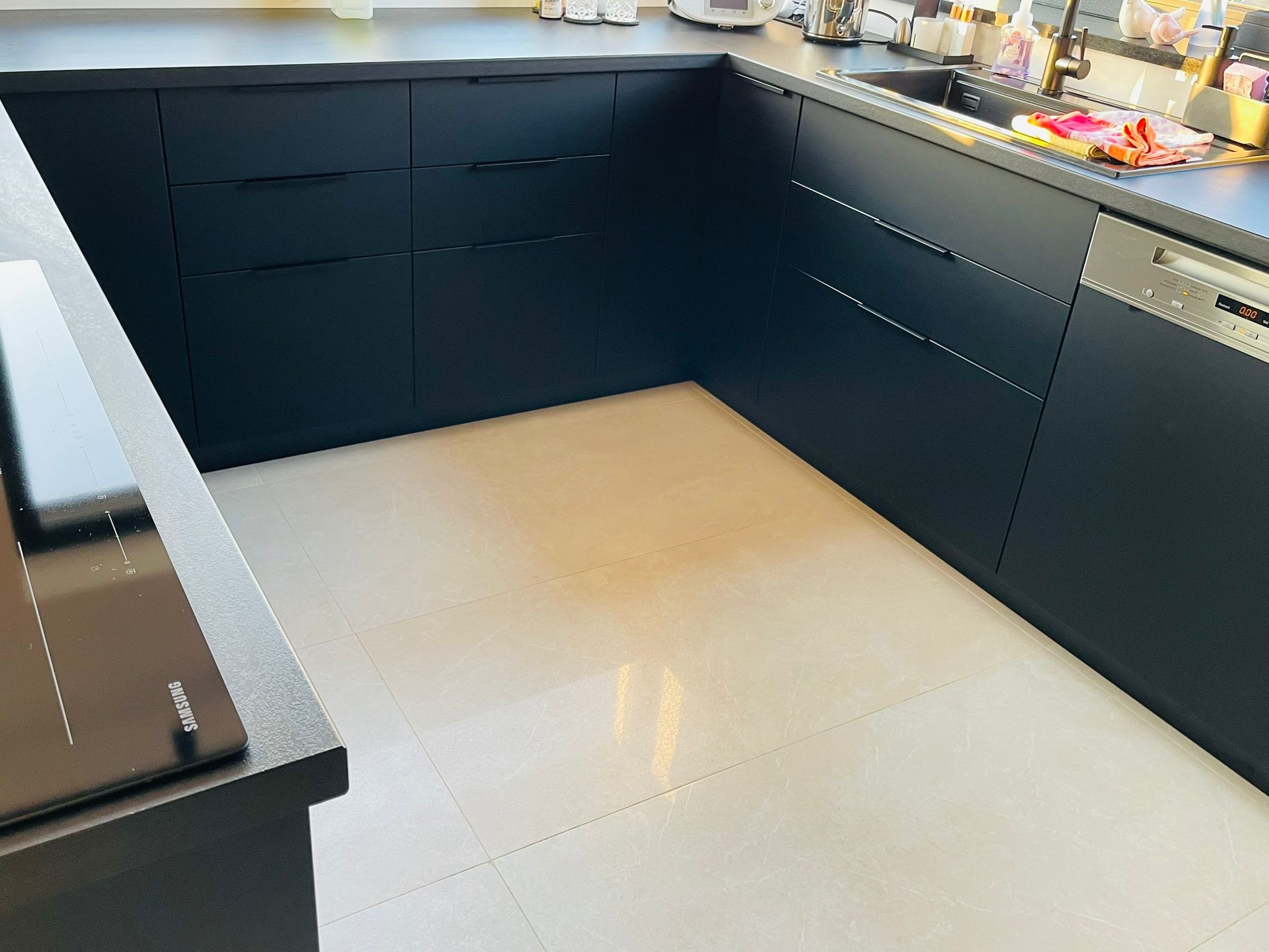 Nowoczesna, narożna kuchnia z matowymi, grafitowymi frontami i beżową podłogą z płytek. Widoczna płyta indukcyjna Samsung i złota armatura przy zlewozmywaku.