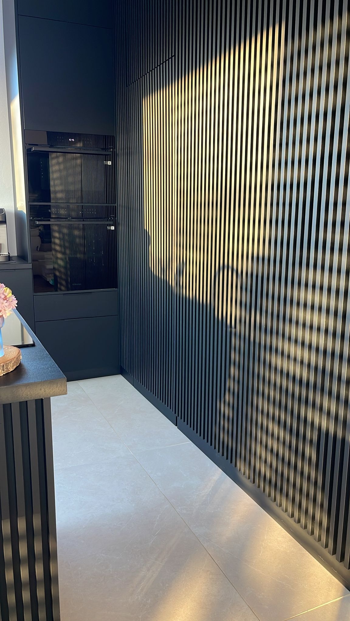 Nowoczesna, czarna kuchnia z pionowymi lamelami na ścianie, piekarnik w zabudowie i jasna podłoga. Minimalistyczny design z efektem światłocienia.