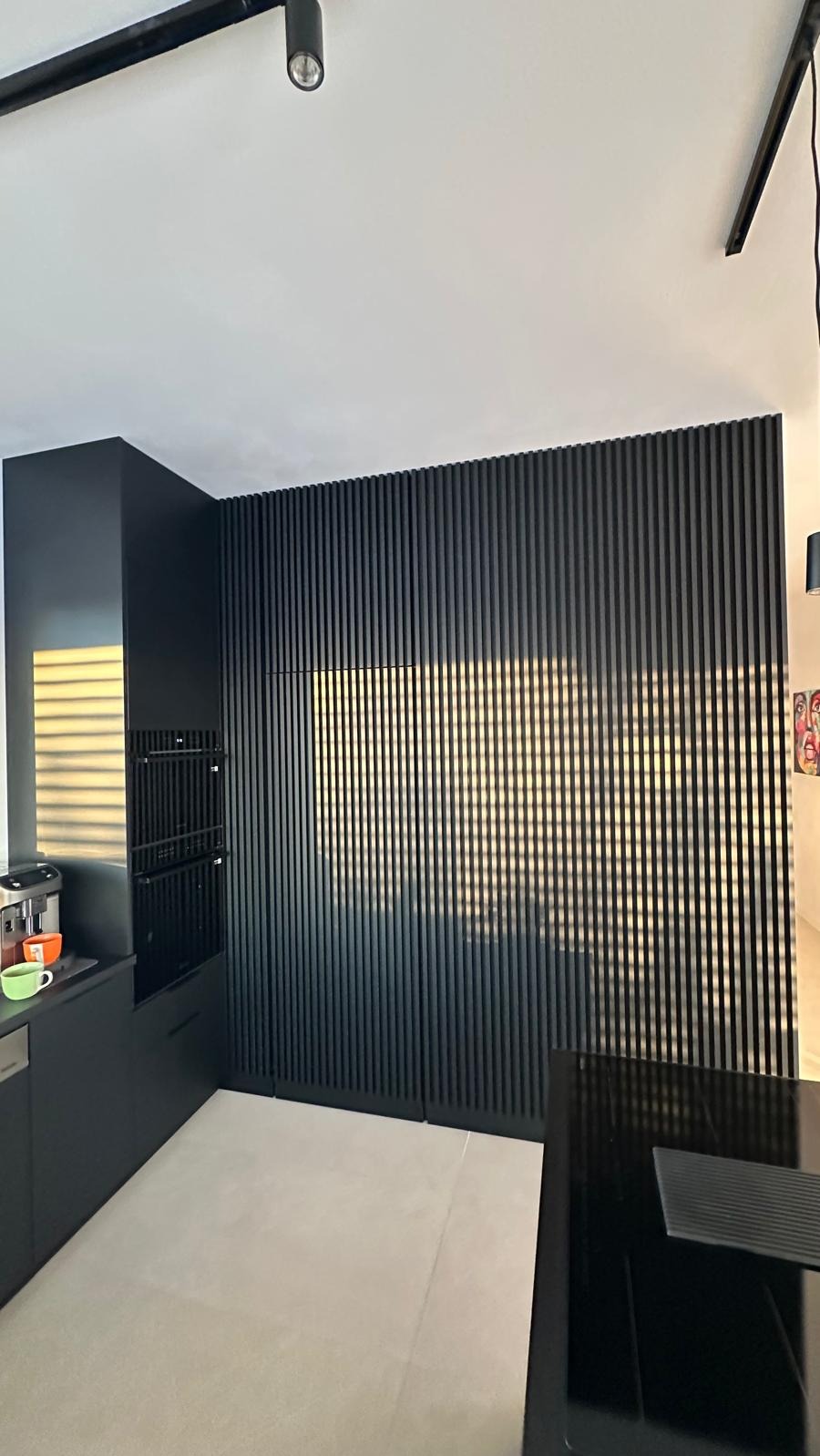 Nowoczesna, czarna kuchnia z lamelami na ścianie, ukrywającymi szafki. Minimalistyczny design, oświetlenie punktowe, jasna podłoga. Widoczny ekspres do kawy.