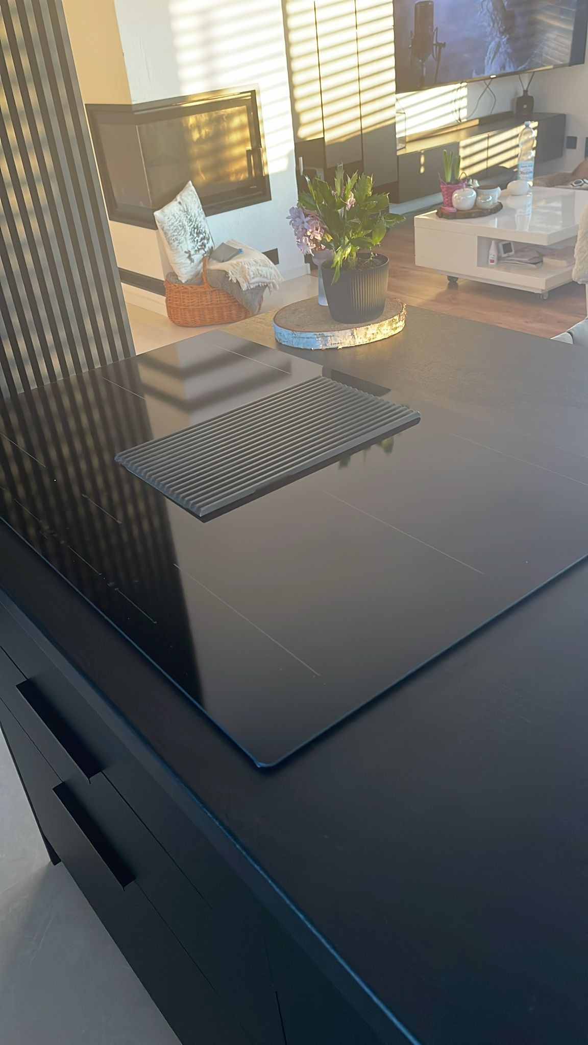 Nowoczesna wyspa kuchenna z czarnym blatem i wbudowaną płytą indukcyjną z pochłaniaczem. W tle salon z kominkiem i telewizorem. Minimalistyczny design.