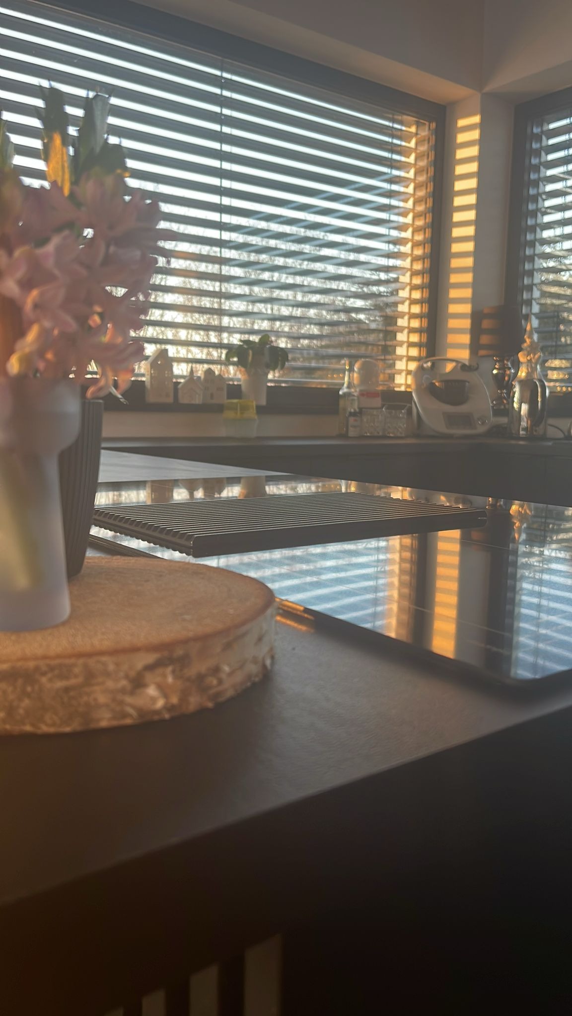 Nowoczesna kuchnia z blatem z ciemnego drewna, na którym stoi ciasto i wazon z kwiatami. W tle okno z żaluzjami przepuszczającymi promienie słoneczne.