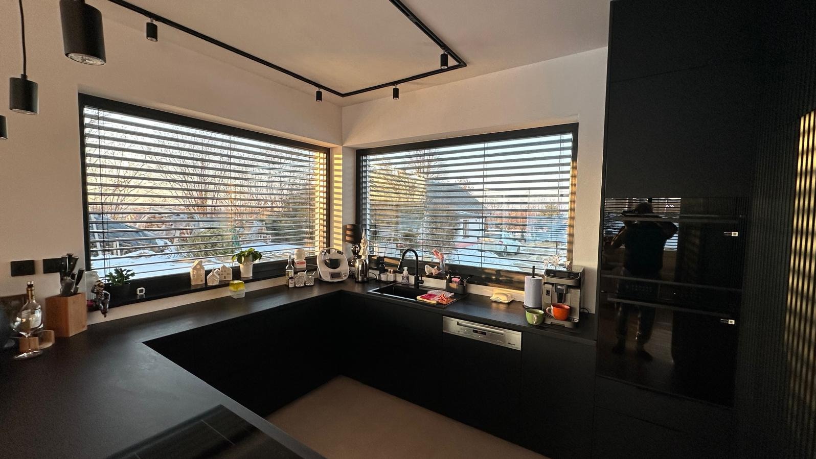 Nowoczesna, czarna kuchnia z dużym oknem i żaluzjami. Minimalistyczny design, oświetlenie punktowe, widok na zimowy krajobraz za oknem.