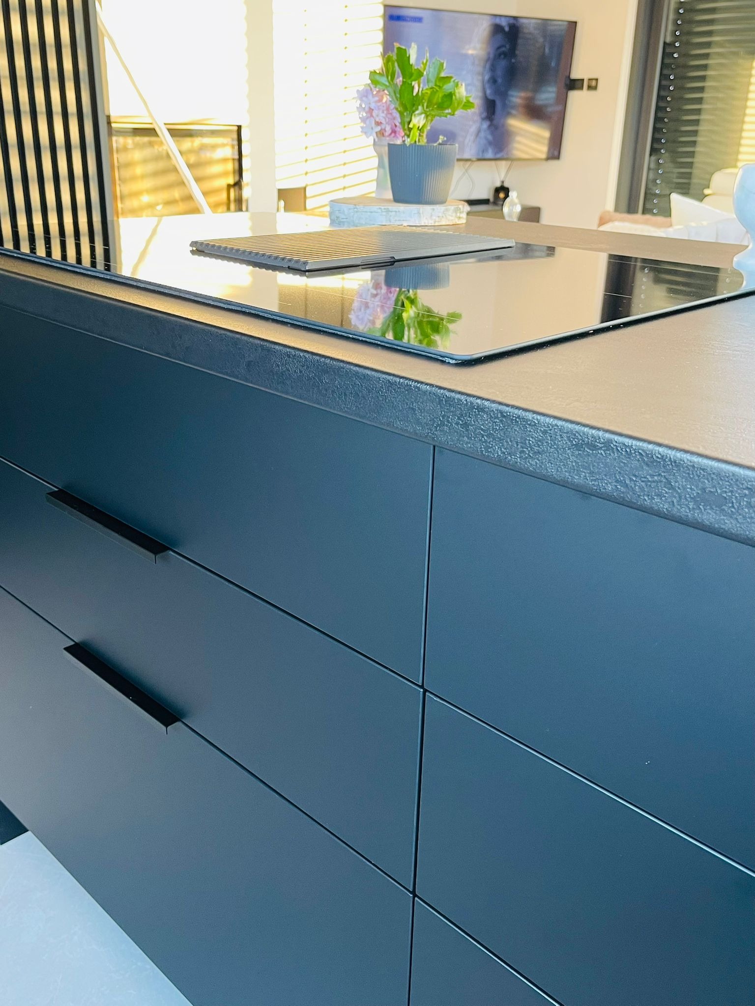 Nowoczesna kuchnia z ciemnymi, matowymi frontami szuflad i blatem z wbudowaną płytą indukcyjną. Na blacie dekoracja z kwiatami w szarej doniczce.