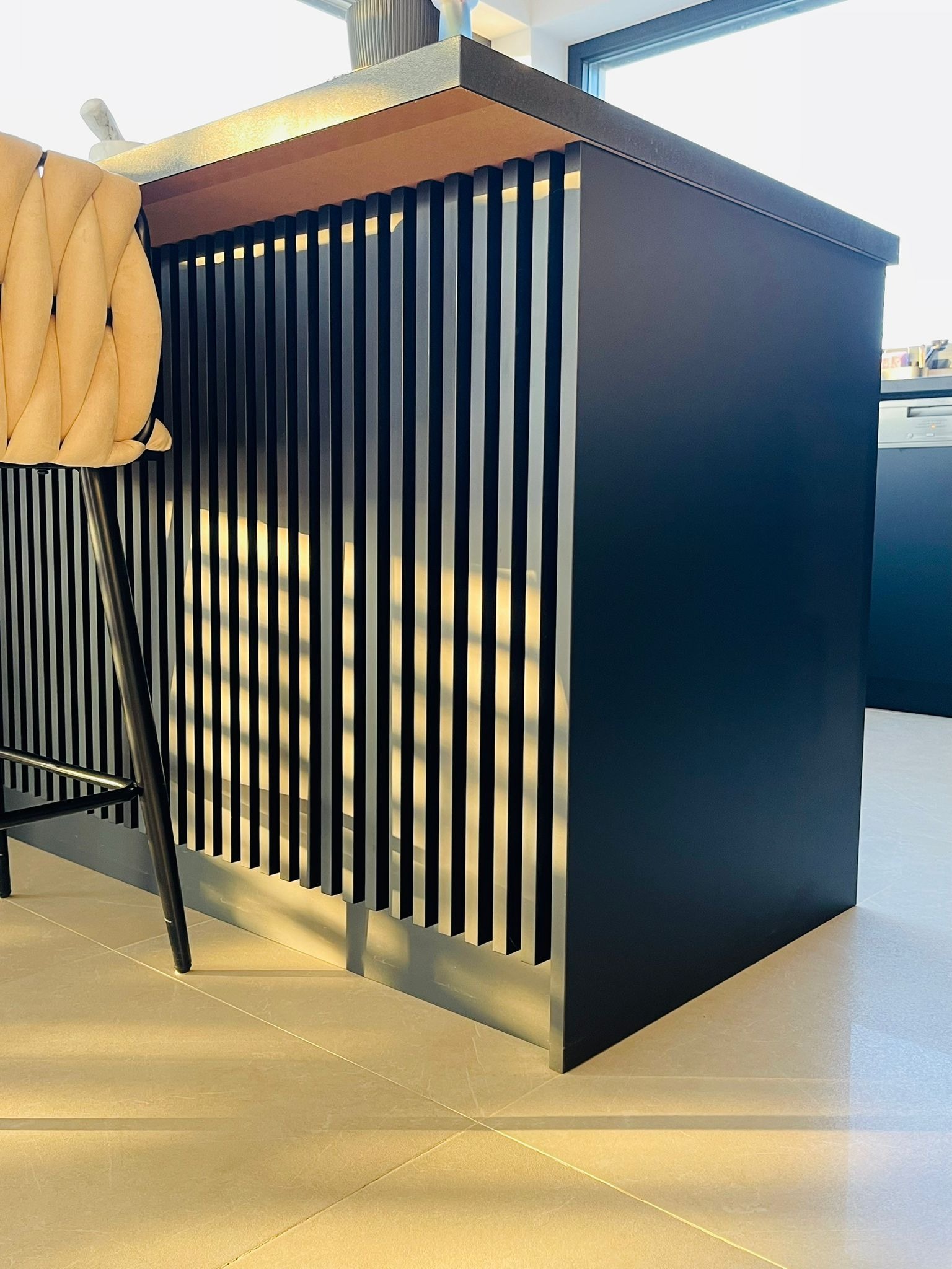 Czarny, nowoczesny barek kuchenny z pionowymi listwami i beżowym blatem, oświetlony promieniami słońca. Obok stoi krzesło barowe z beżowym siedziskiem.