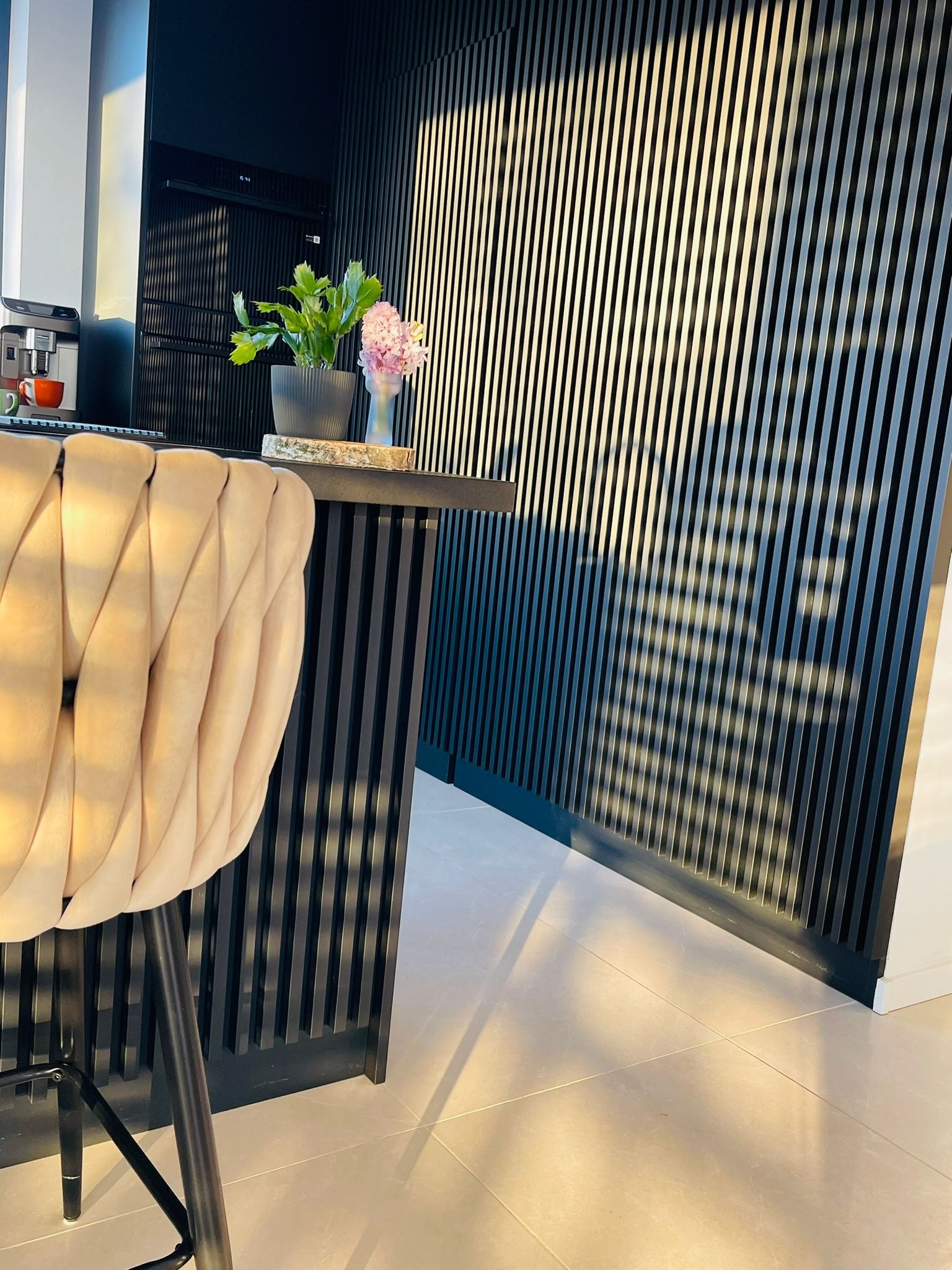 Nowoczesna kuchnia z czarnymi lamelami na ścianie i wyspą z beżowym krzesłem. Na blacie kwiaty i ekspres do kawy. Minimalistyczny design, ciepłe oświetlenie.