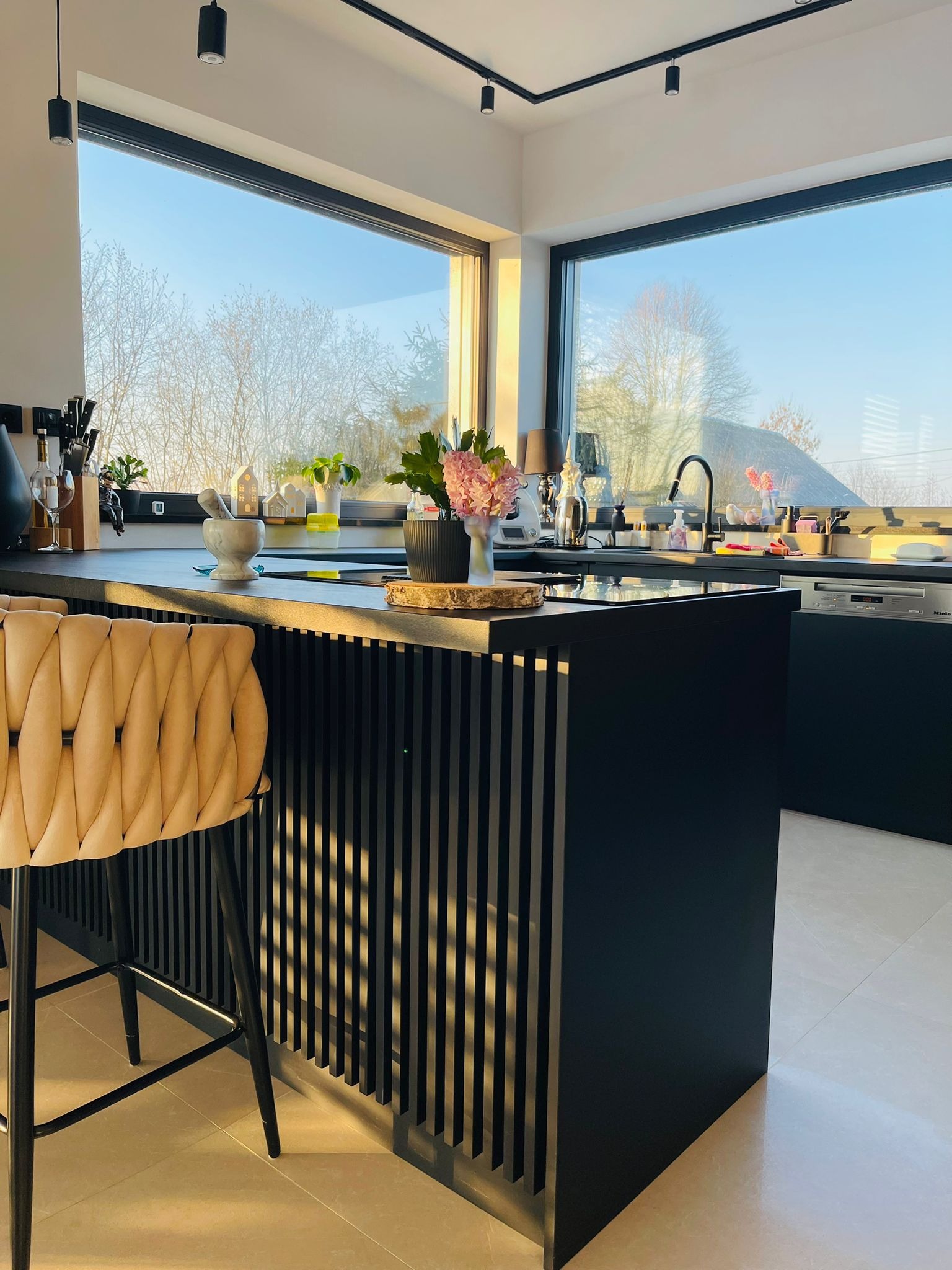 Nowoczesna kuchnia z czarną wyspą i beżowym krzesłem barowym, oświetlona naturalnym światłem z dużego okna. Minimalistyczny design z akcentami roślinnymi i dekoracyjnymi.