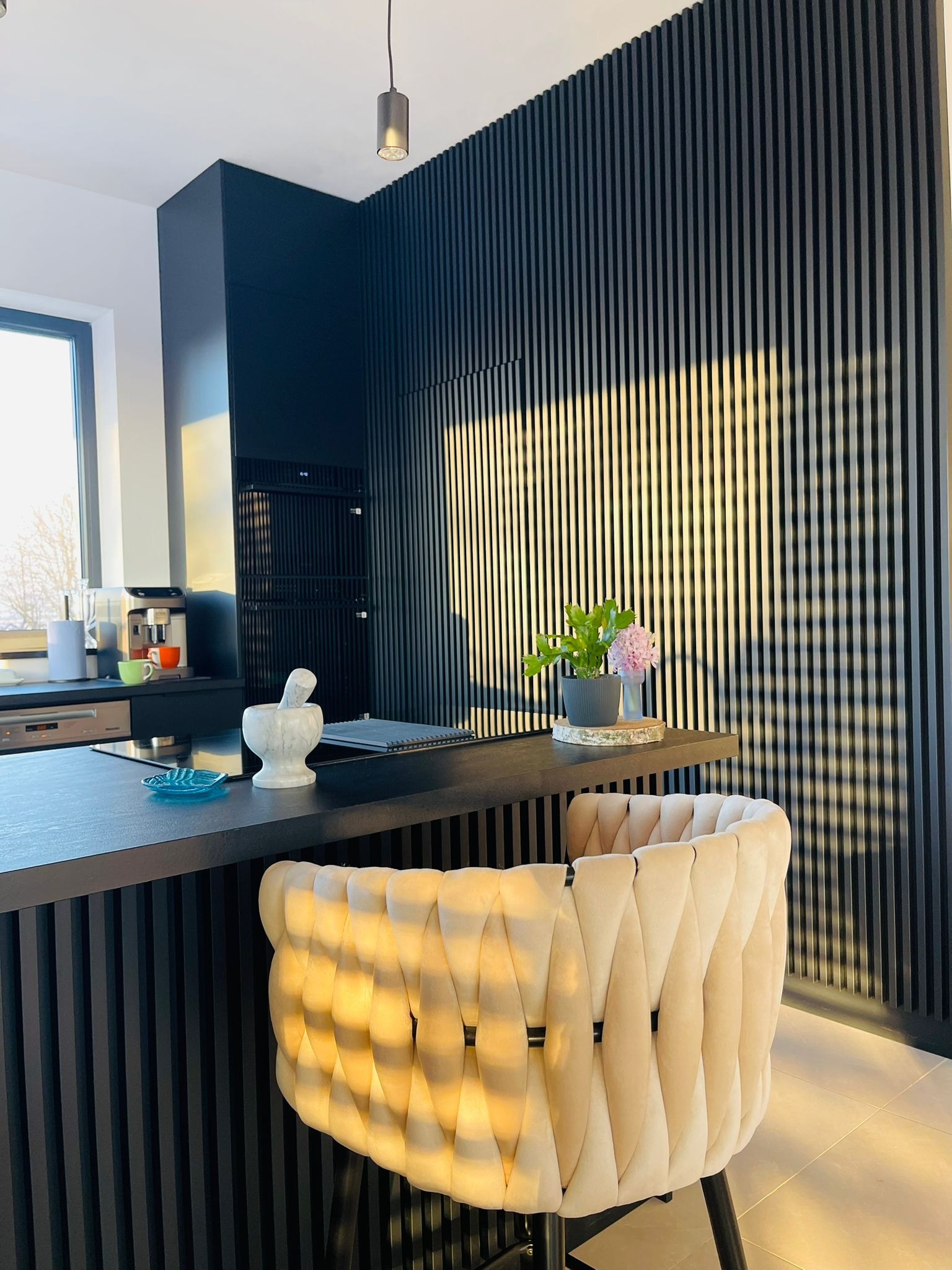 Nowoczesna kuchnia z czarnymi lamelami na ścianie, marmurowym moździerzem na blacie i beżowym krzesłem barowym. Minimalistyczny design z akcentami roślinnymi.