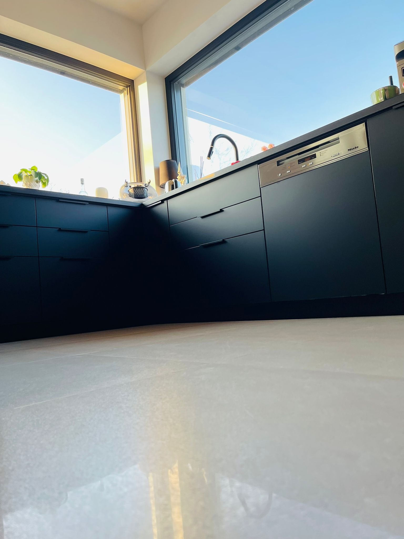 Nowoczesna, czarna zabudowa kuchenna z dużym oknem i widoczną zmywarką Miele. Ujęcie z dołu podkreśla minimalistyczny design i jasną podłogę.