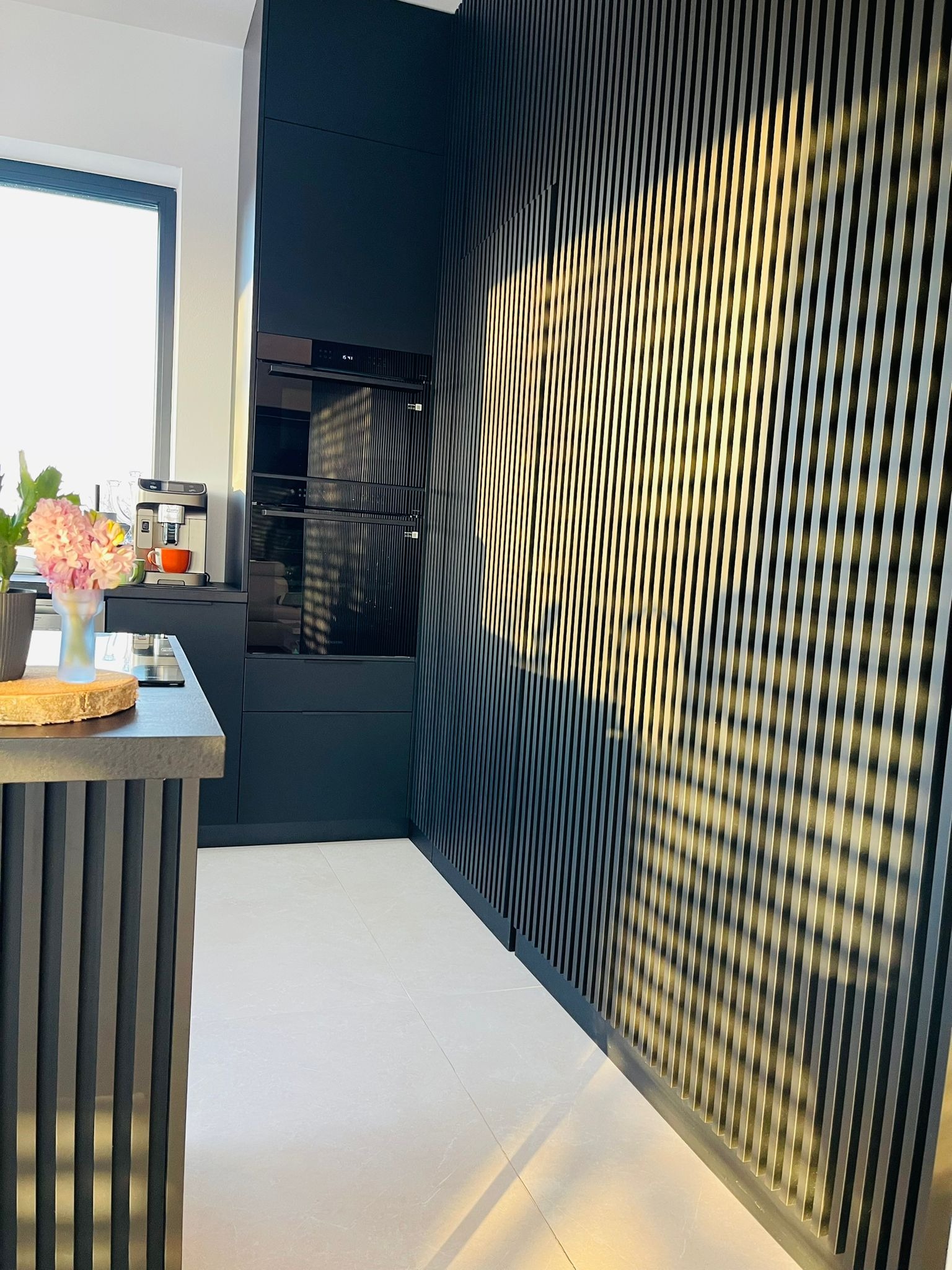 Nowoczesna kuchnia w odcieniach grafitu z dekoracyjną ścianą z pionowych listew, oświetlonych promieniami słońca. Minimalistyczny design, eleganckie wykończenie.