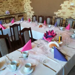 Uroczysta dekoracja stołu z białym obrusem, różowymi i fioletowymi serwetkami, zastawą stołową i kwiatową dekoracją w centrum; kamienna ściana w tle.