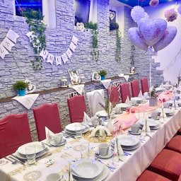 AgawaHouse - Uroczysty stół komunijny z dekoracjami: balony w kształcie serc, girlanda z napisem 'I Komunia', elegancka zastawa i kwiatowe akcenty. Kamienna ściana w tle.