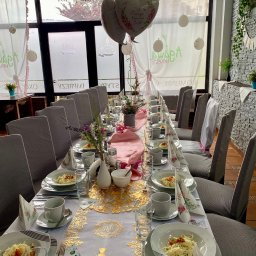 AgawaHouse - Uroczysty stół komunijny: biały obrus ze złotymi zdobieniami, zastawa, kwiaty, balony. Dekoracja okna z napisem 'Święta Komunia'. Krzesła w szarych pokrowcach.