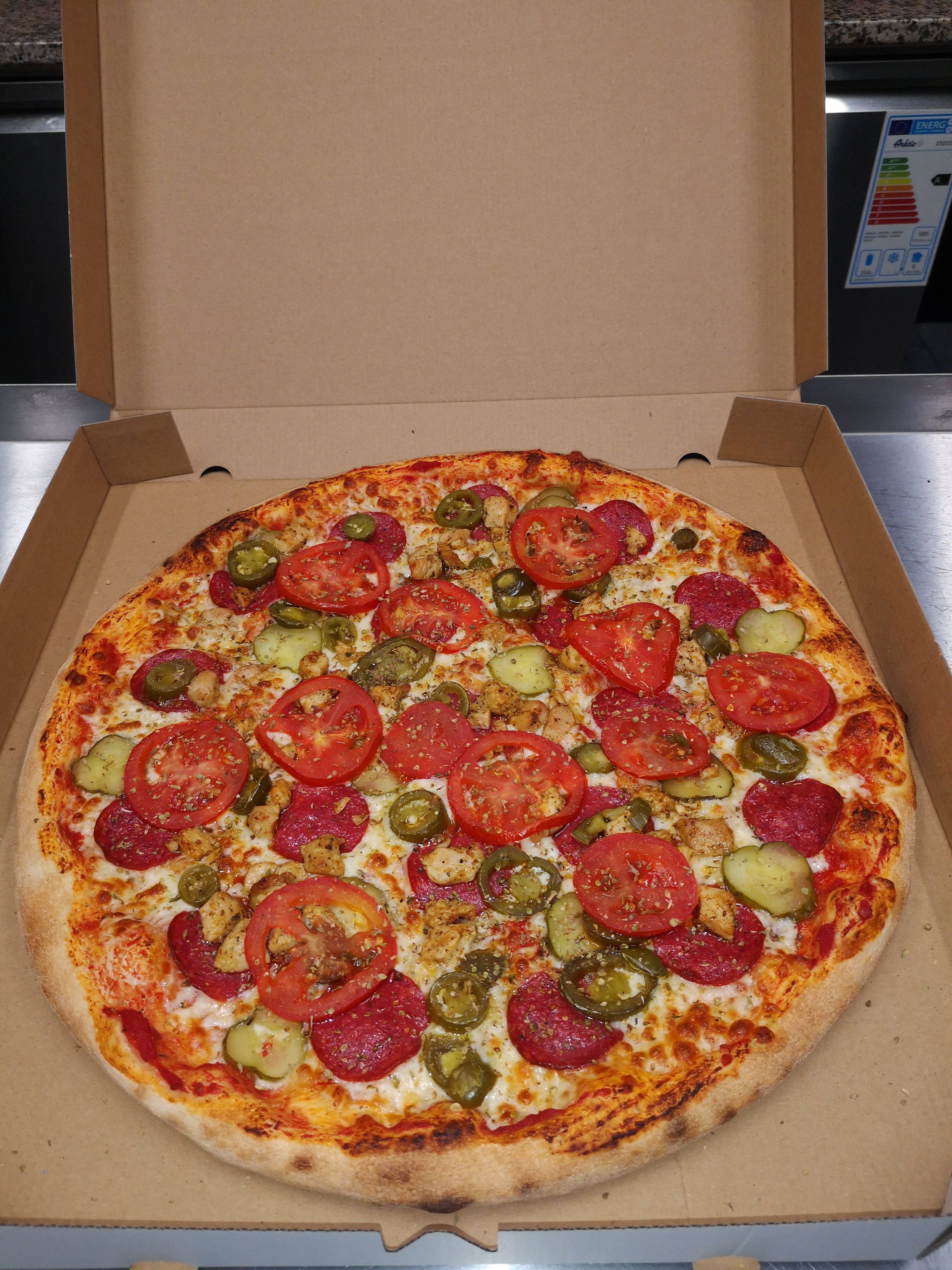 Pizza w kartonie z pepperoni, pomidorami, kurczakiem i papryczkami jalapeno, posypana ziołami, z widoczną etykietą energetyczną na lodówce w tle.