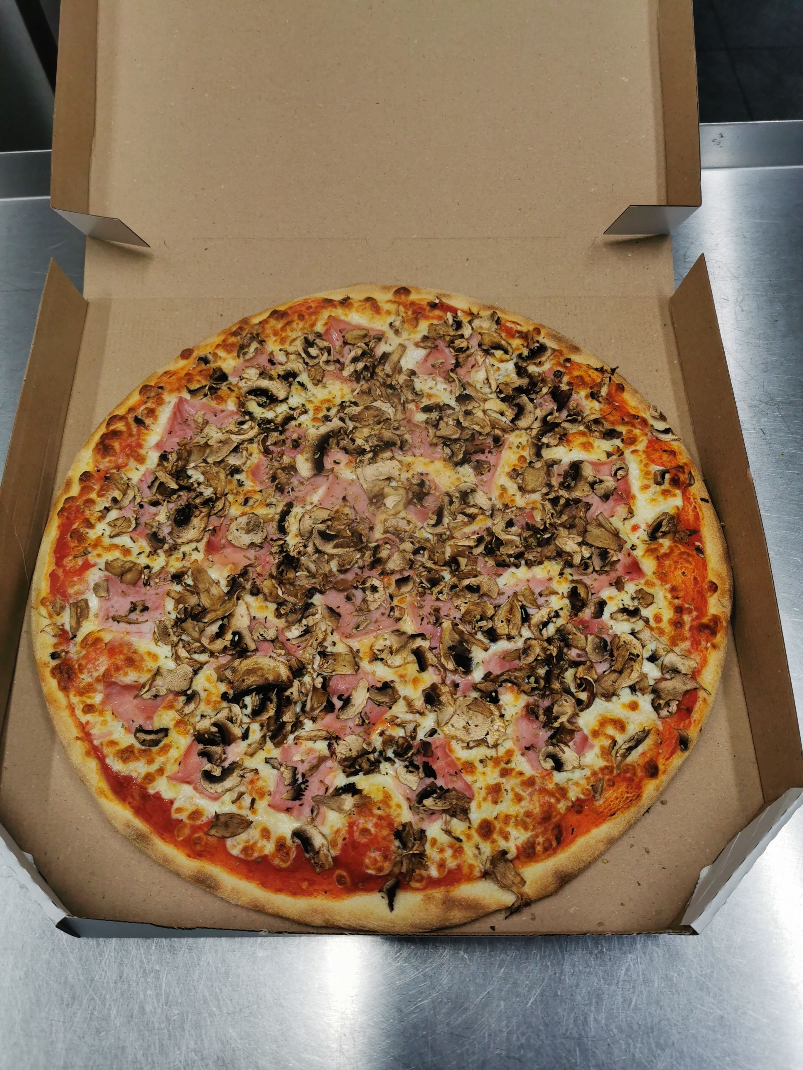 Okrągła pizza z pieczarkami i szynką, umieszczona w otwartym kartonowym pudełku na metalowym blacie.