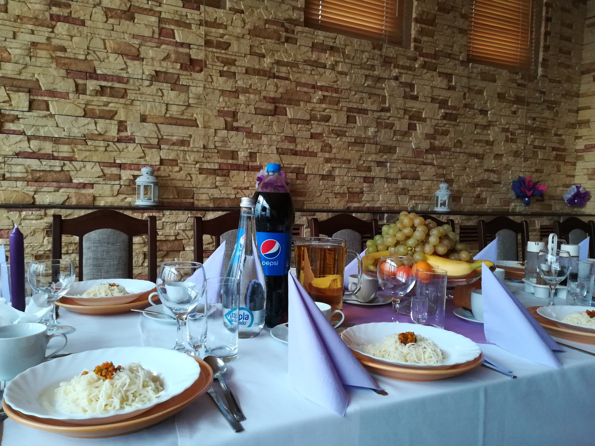 Uroczysty stół z białym obrusem, nakryty zastawą, daniami z makaronu z pomarańczową posypką, szklankami, napojami w butelkach i dzbanku, owocami, fioletowymi serwetkami, na tle ściany z cegieł...