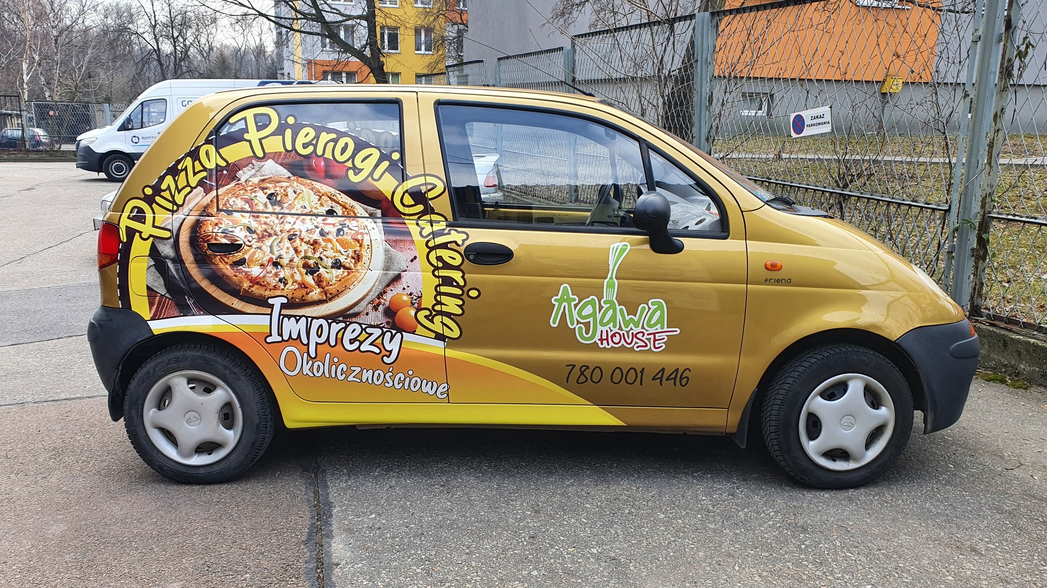 Złoty samochód z reklamą pizzy, pierogów i cateringu na boku, zaparkowany na ulicy obok ogrodzenia.