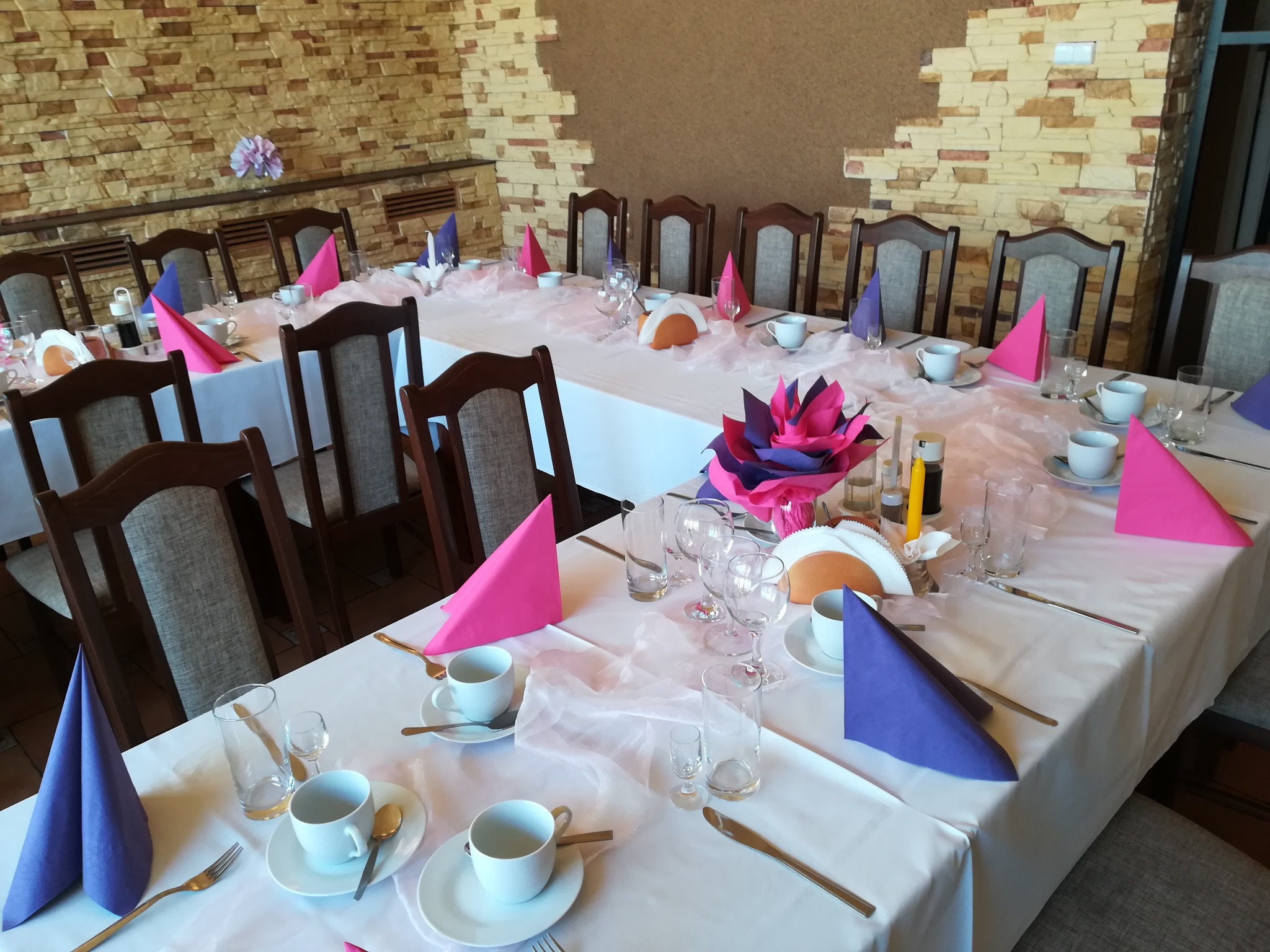 Uroczysta dekoracja stołu z białym obrusem, różowymi i fioletowymi serwetkami, zastawą stołową i kwiatową dekoracją w centrum; kamienna ściana w tle.