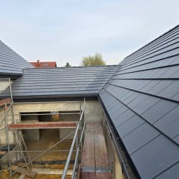 Ostafiński Mariusz Usługi Dekarsko - Ciesielsko - Blacharskie - Nowy dach z szarej dachówki ceramicznej, widok z góry na budynek w trakcie budowy. Na pierwszym planie rusztowanie i fragment niezadaszonej konstrukcji.