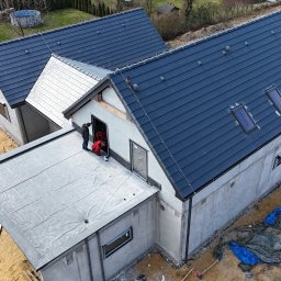 Ostafiński Mariusz Usługi Dekarsko - Ciesielsko - Blacharskie - Nowoczesny dom w trakcie budowy z ciemną dachówką i oknami dachowymi. Widoczni pracownicy montujący elementy. Ujęcie z góry ukazuje postęp prac.