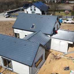 Ostafiński Mariusz Usługi Dekarsko - Ciesielsko - Blacharskie - Widok z góry na dom w trakcie budowy z szarym dachem z dachówki. Na dachu pracują dwie osoby. W tle inne budynki i drzewa. Widoczne materiały budowlane.